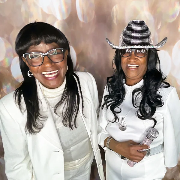 two women wearing white smiling at backyard party photo booth in Carson California with glitter microphone prop